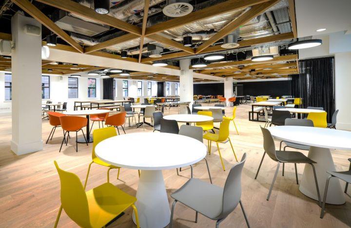 Large room with exposed industrial ceiling with informal seating area with white tables and orange, grey and yellow chairs.