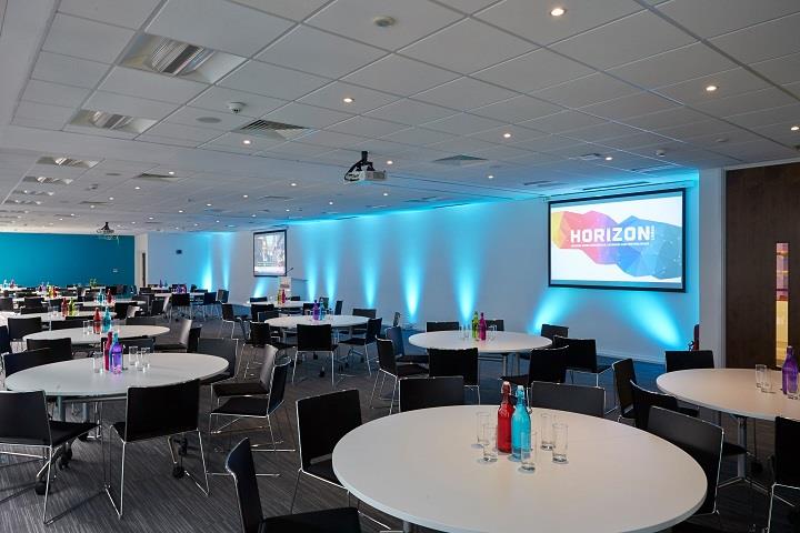 Meeting room with blue feature wall and uplighters with wound tables and black chairs.